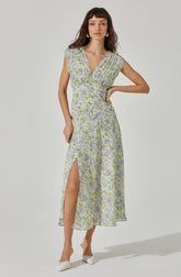A woman is wearing a long, flowy dress with a floral pattern in shades of purple, green, and white. The dress has a V-neckline and short, slightly caped sleeves. It features a wrapped bodice and a high slit on the left side, revealing her leg. The fabric appears lightweight and semi-transparent. She is also wearing white shoes with a bow detail.